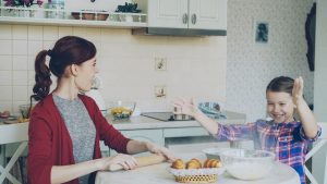 Young mother and cute daughter cooking together talking in the kitchen on weekend. Little funny girl clapping hands with flour and laughing. Family, food, home and people concept